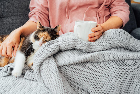 Woman enjoying a hot beverage on couch while cuddling with a cat and blanket.