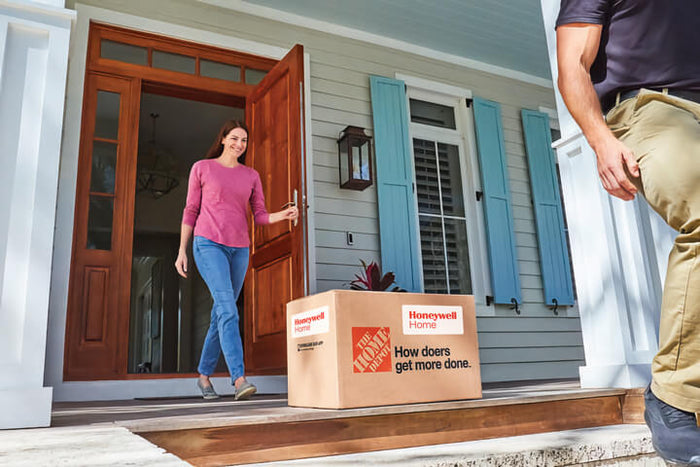 A woman receives a package on her porch. The package has the Home Depot logo and the Honeywell Home logo along with text that reads: How doers get more done.
